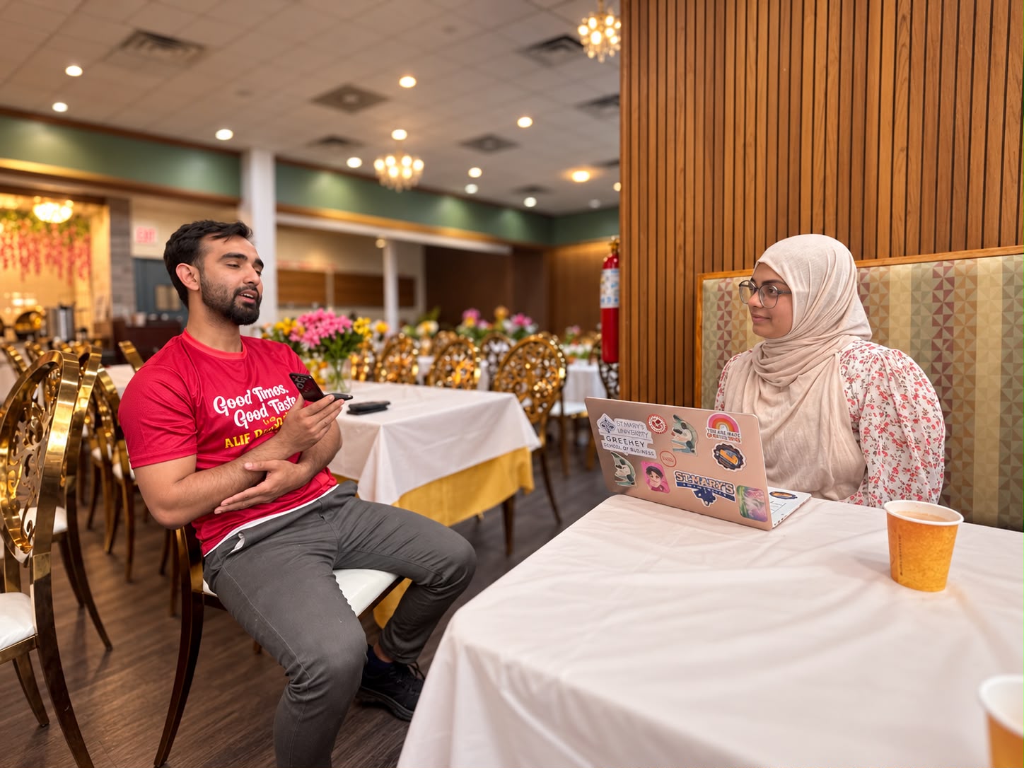 Hasnat bhai in a red shirt sits across from a researcher with a laptop in a decorated restaurant